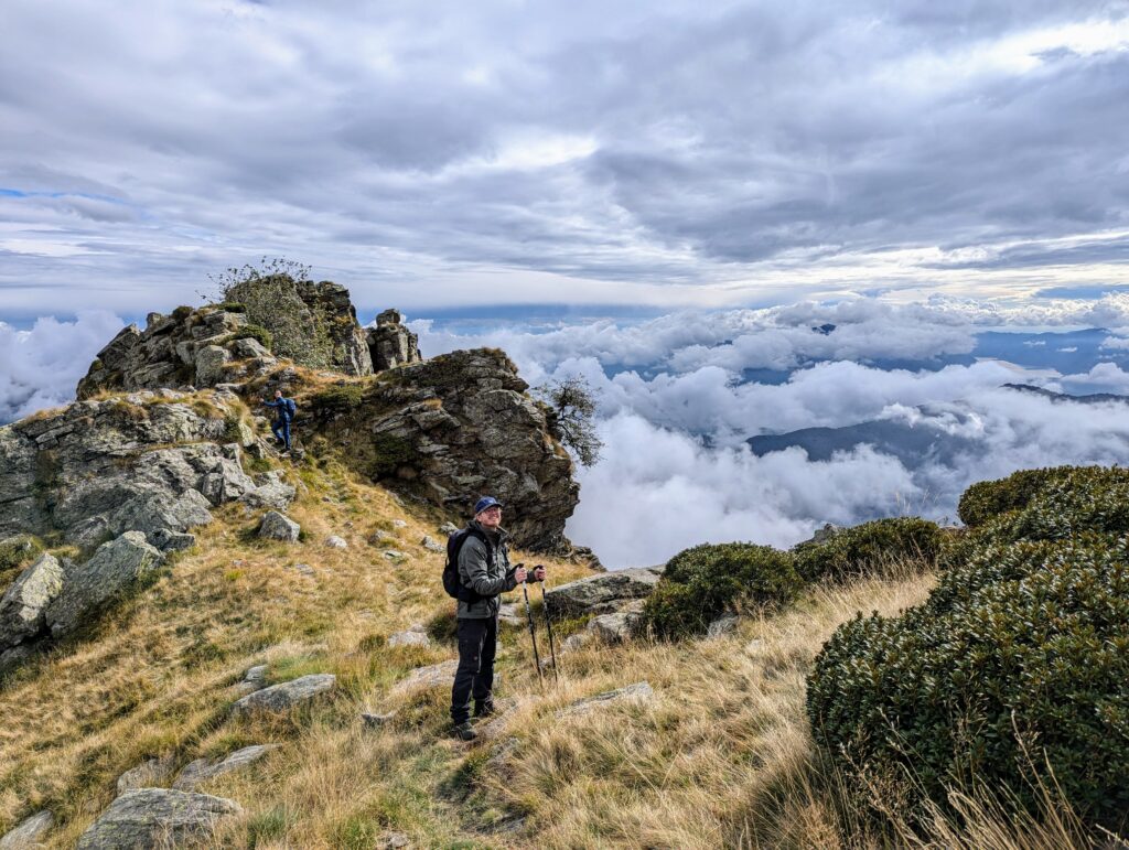 Deelnemers navigeren over een rotsachtig pad op grote hoogte, omringd door grasland en dramatische wolkenformaties die de omliggende bergen bedekken.