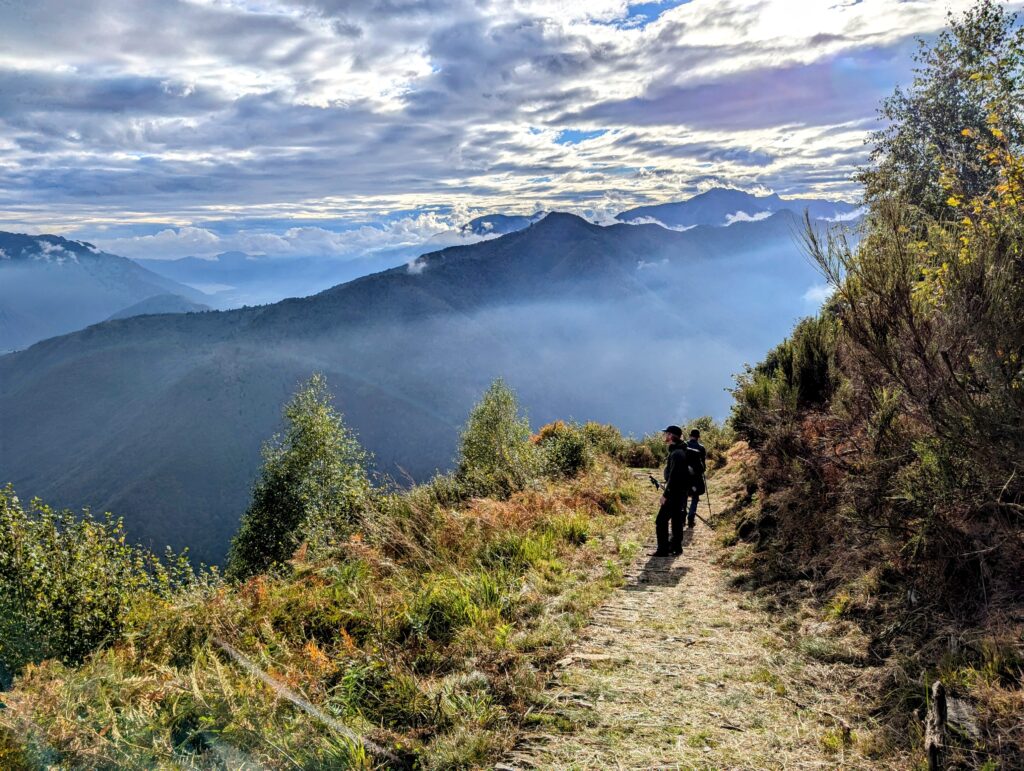 Twee deelnemers lopen op een smal pad door een bergachtig landschap, omgeven door groen en met een spectaculair uitzicht op valleien en bergtoppen onder een deels bewolkte hemel.